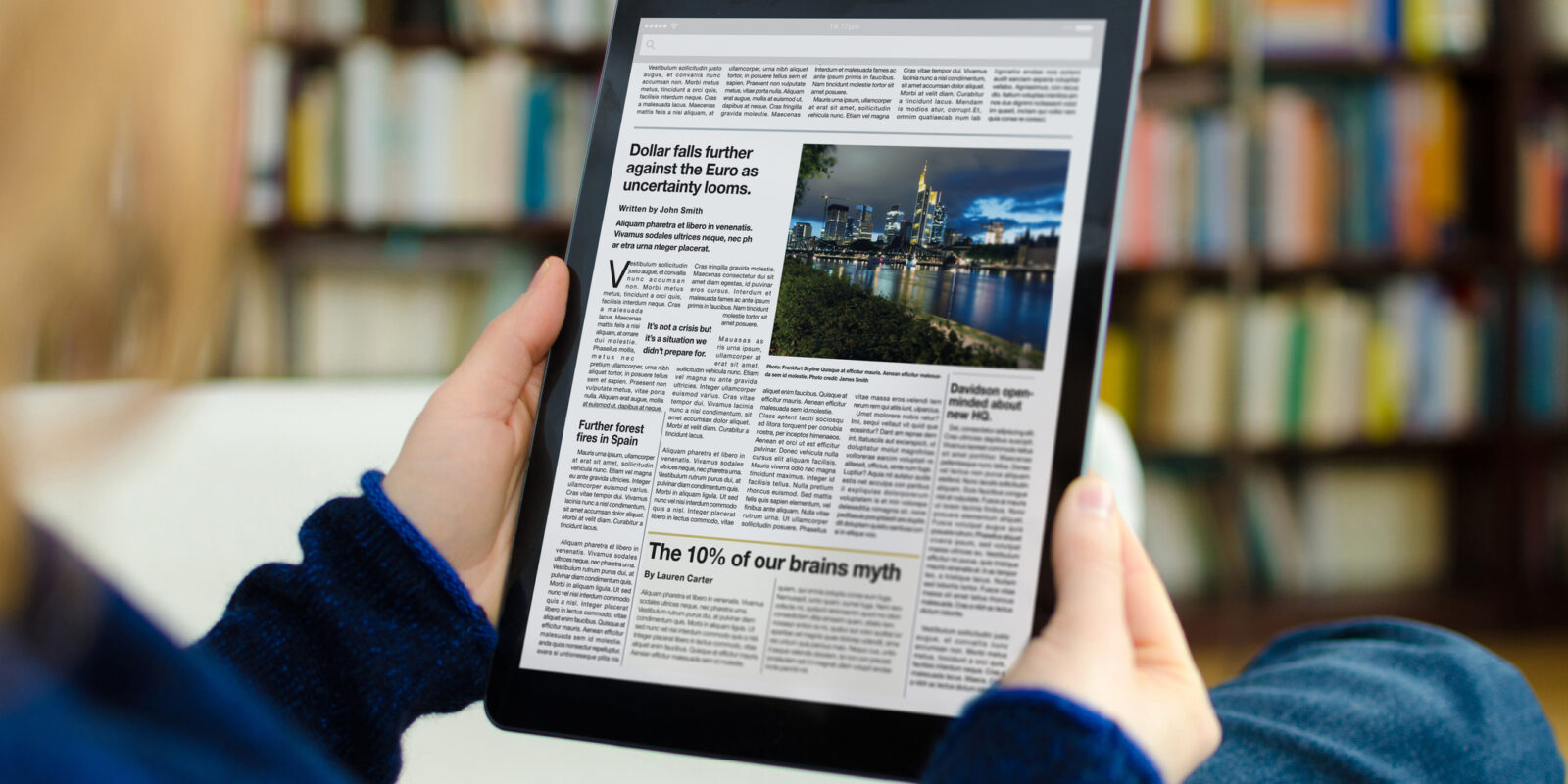 A digital newspaper on a modern tablet computer held by a young woman, who is reading the day’s news. The photo was taken by looking over the model's shoulder. A bookshelf is visible in the background of the picture. The newspaper on the device screen was created by myself in Indesign, I own the copy right of the design and layout. The image of Frankfurt on the device is my own, I own the copy right. The text is all lorem ipsum except for the titles, which are generic and entirely fictional – these were written by myself.